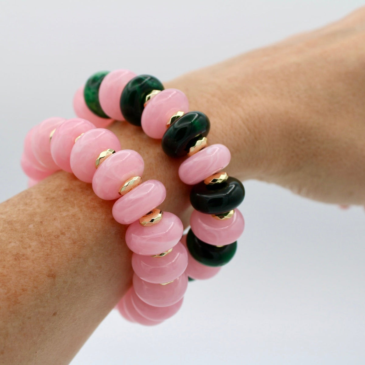 Two beaded stretch bracelets on a person's wrist, featuring pink and dark green disc-shaped beads separated by small gold spacer rings, against a plain white background.