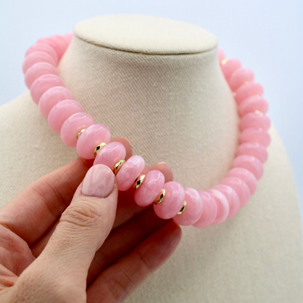 A pink beaded necklace with small gold spacer rings displayed on a cream jewelry bust, with a hand gently holding the front of the necklace.